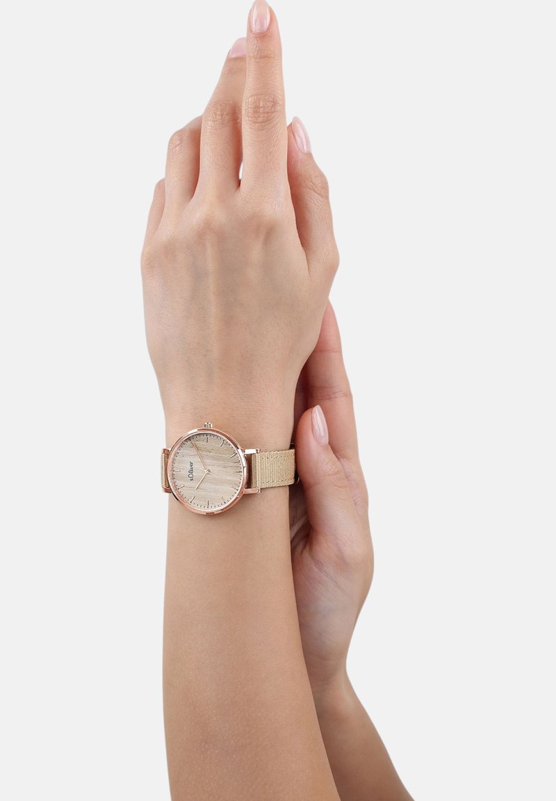 Persoon met een beige stoffen polsbandhorloge met een licht houten wijzerplaat, die zachtjes zijn pols vasthoudt met de andere hand.