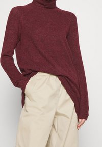 Burgundy turtleneck sweater with a textured knit, loose fit, and side slits, paired with light beige relaxed-fit trousers.