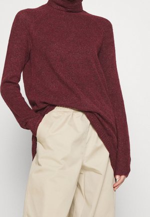 Un pull col roulé bordeaux avec un tricot texturé, coupe ample et fentes sur les côtés, associé à un pantalon beige clair à coupe décontractée.