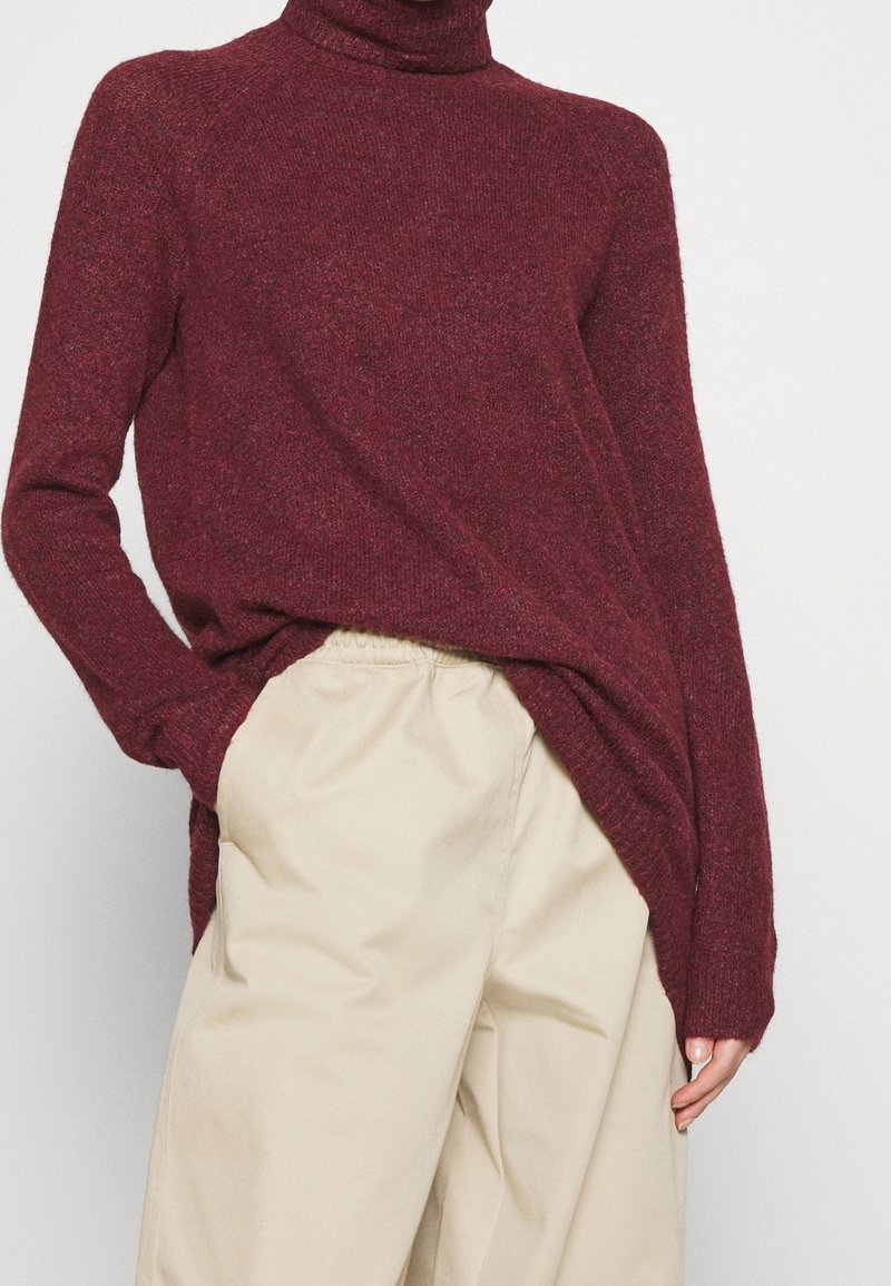 Burgundy turtleneck sweater with a textured knit, loose fit, and side slits, paired with light beige relaxed-fit trousers.