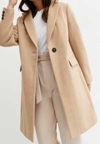 Manteau beige en mélange de laine avec une coupe cintrée, de larges revers et une fermeture à un bouton. Dispose de poches latérales et d'une texture lisse et douce.