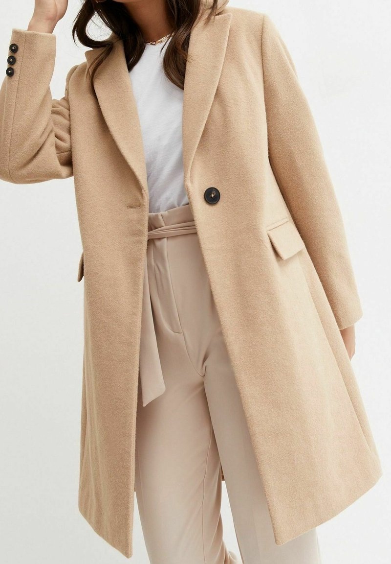 Manteau beige en mélange de laine avec une coupe cintrée, de larges revers et une fermeture à un bouton. Dispose de poches latérales et d'une texture lisse et douce.