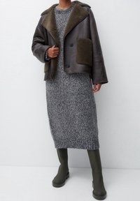 Chaqueta de cuero forrada de piel de oveja marrón con dos bolsillos frontales, sobre un vestido de punto largo gris y botas altas de ante verdes oscuras.