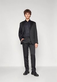 Crni odijelo s uzorkom, teksturirani blejzer i hlače, u kombinaciji s crnom košuljom. Obuća uključuje crne polirane cipele.