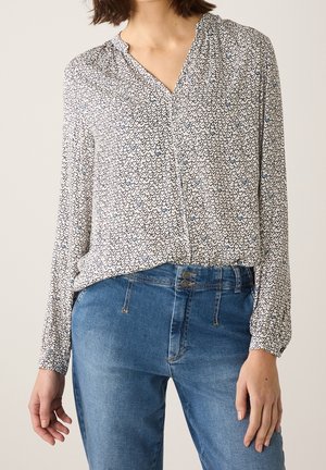 Vrouw die een wit blouse met lange mouwen draagt met kleine zwarte en blauwe hartjespatronen, ingestopt in een blauwe spijkerbroek.