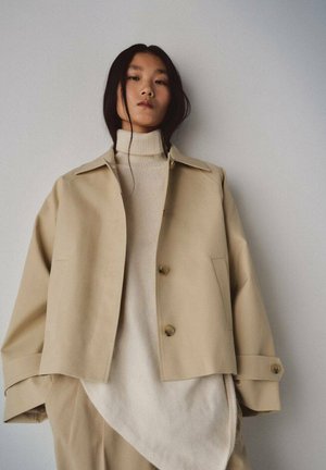Woman with long black hair wearing beige oversized jacket and light beige turtleneck sweater, standing against plain light wall.