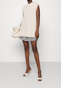 Vestido de punto en beige, sin mangas con escote redondo. Combinado con una falda corta negra con flores. Se acompaña de mules blancos de tacón y un clutch acolchado.