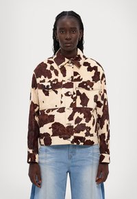 Jeansjacka med ko-print som har en beige botten med mörkbruna fläckar, knäppning fram och två bröstfickor. Croppad design med långa ärmar.
