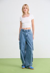 T-shirt bianco a coste, accorciato, abbinato a pantaloni cargo in denim blu a gamba larga con tasche laterali, completato da sandali blu su una superficie verde.
