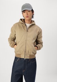 Sweatshirt bege com fecho na frente, bolso frontal, punhos e bainha canelados. Combinado com jeans escuros. O boné é cinza claro com a aba curva.