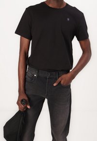 Camiseta de algodón negra con cuello redondo y mangas cortas, combinada con jeans de mezclilla gris oscuro. Mano sosteniendo un bolso negro de diseño minimalista.