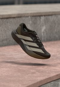 Καφέ αθλητικό παπούτσι με υφή στο επάνω μέρος, που διαθέτει φαρδιές λευκές ρίγες, μαύρα κορδόνια και λείο πέλμα, εμφανιζόμενο σε αιωρούμενη θέση.