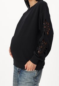 Schwarzer Sweatshirt mit ausgeschnittenem Blumenmuster an den Ärmeln, Rundhalsausschnitt und gerippten Bündchen; kombiniert mit hellblauen Jeans.