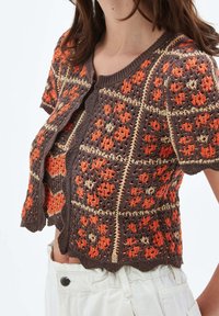 Cárdigan de manga corta de ganchillo en patrones florales geométricos en marrón, naranja y beige, combinado con pantalones blancos de cintura alta.