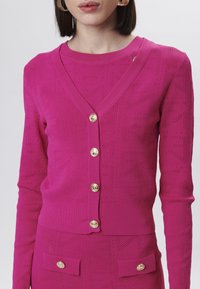 Cardigan a maglia fucsia con scollo a V, bottoni dorati e un motivo geometrico con perforazioni. Texture liscia e design aderente.