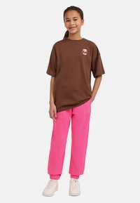 Brun bomullströja med korta ärmar och vit broderad detalj, i kombination med rosa sweatpants med mudd, bärs med vita sneakers.