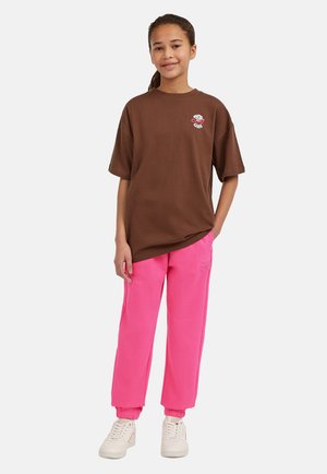 Maglietta di cotone marrone a maniche corte con dettagli ricamati bianchi, abbinata a pantaloni della felpa rosa con orli, indossati con scarpe da ginnastica bianche.