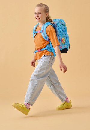 Smiling girl walking wearing a bright blue backpack with stars, orange T-shirt, torn light jeans, pink socks, and yellow sneakers on beige background.