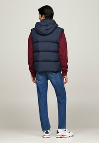 Navy puffer vest with hood, worn over a burgundy sweatshirt. Paired with blue jeans and white sneakers featuring red and navy accents.