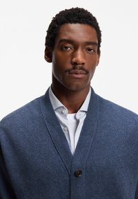 Blå kofta med v-ringning, ribbad struktur och en svart knapp som stängning, bärs över en vit skjorta med knappar och texturerat mönster.