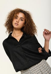 Black button-up shirt made from soft fabric, featuring a collar and rolled sleeves. Paired with a checked skirt, showcasing contrasting patterns.