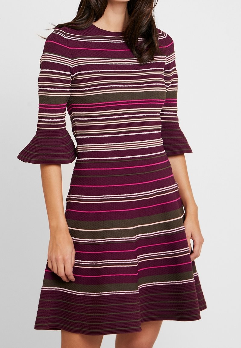 Femme portant une robe rayée violette, rose et marron avec des manches évasées trois-quarts et une jupe évasée.