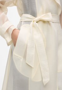 Chemise en tissu léger et transparent de couleur crème douce. Elle présente une ceinture nouée et un détail de poche, avec une couture délicate sur la ceinture.