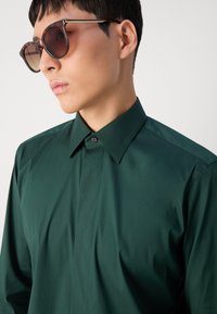 Camisa de botón verde oscuro con un cuello estructurado, textura suave y un ligero brillo. Gafas de sol grandes y redondas con un tinte degradado.
