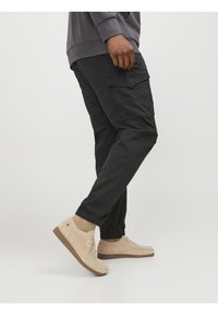 Zwarte cargobroek met elastische boorden, voorzien van meerdere zijzakken. Draagt men met lichtbeige suède schoenen met een gestructureerde zool.
