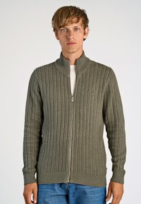 Green knitted zip-up cardigan with a high collar, featuring a ribbed texture, long sleeves, and fitted cuffs. Worn over a white shirt.