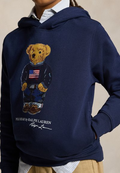 Laivastonsininen huppari, jossa karhu pukeutuneena villapaitaan, jossa on Amerikan lippu, maastohousut ja ruskeat kengät, teksti "Polo Bear by Ralph Lauren".