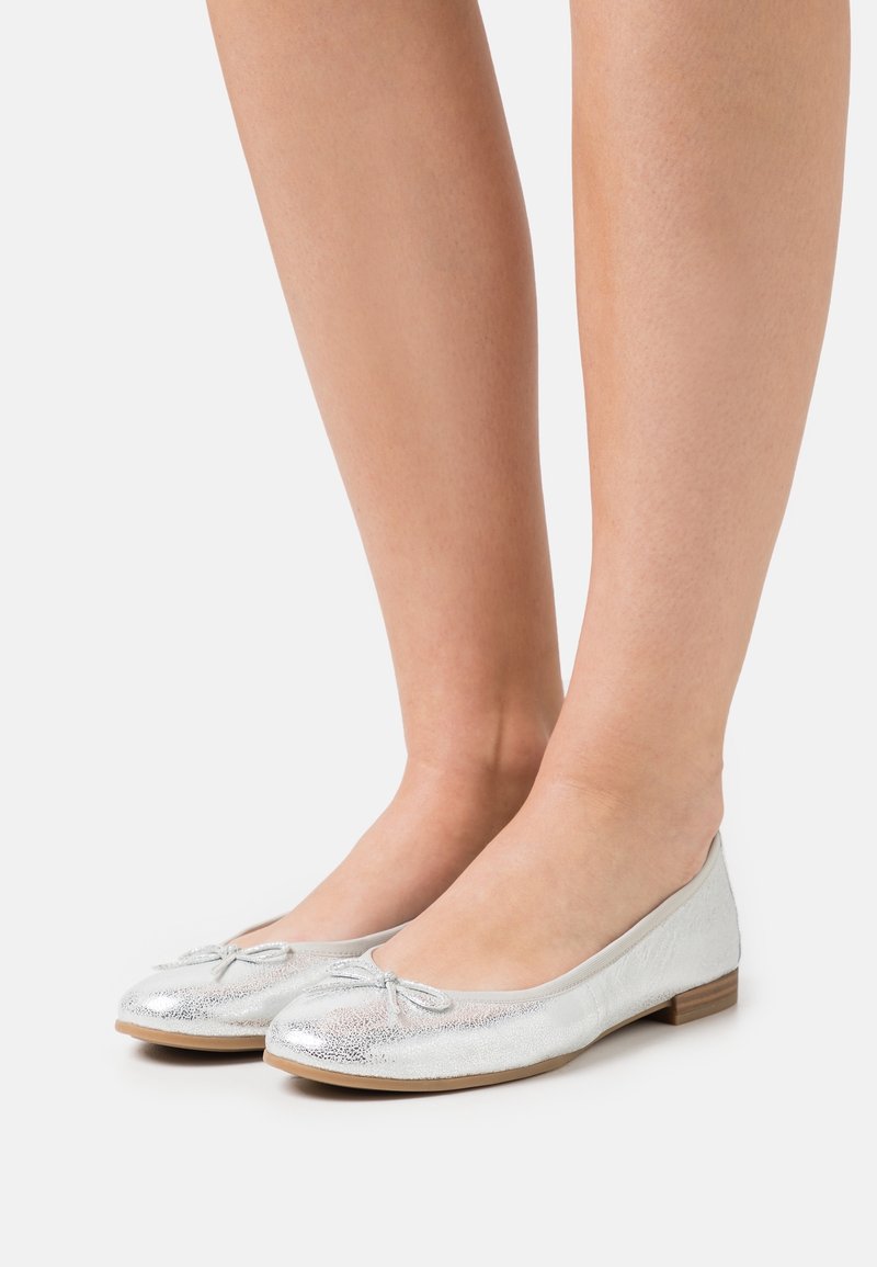 Pieds portant des ballerines argentées avec de petits nœuds, présentés sur un fond clair uni.