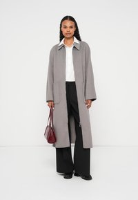 Grå lang frakke med blød tekstur, sidelommer og en klassisk krave, kombineret med en hvid skjorte og sorte bukser; holder en bordeaux håndtaske.