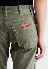 Olívazöld kordbársony nadrágon barna bőr folt található, rajta a "Wrangler" logóval, függőleges bordázott textúrával és klasszikus hátsó zseb kialakítással.