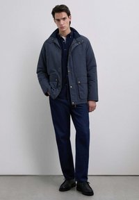 Nuori mies, joka on pukeutunut tummansiniseen takkiin tumman paidan ja vastaavien tummien housujen kanssa, sekä mustiin kenkiin, seisoo yksinkertaista vaaleaa seinää vasten.