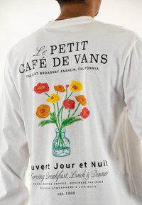 Camisa blanca de manga larga con un diseño floral colorido en la parte posterior, con texto sobre un café en Anaheim. Nota el tarro y la diversidad de colores de las flores.