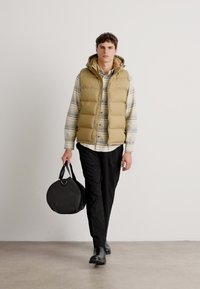 Gilet imbottito khaki sopra una camicia a righe con bottoni, pantaloni neri e stivaletti. La modella porta una borsa rotonda nera. Sfondo semplice.