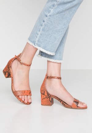 Orange snake-print heeled sandals with a square toe, ankle strap, and block heel. The texture is smooth with a glossy finish.