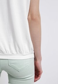 Personne portant un haut blanc sans manches et un pantalon vert clair, vue de dos se concentrant sur le bas du torse et le bras droit.