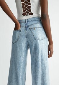 Vaaleansiniset leveälahkeiset farkut, joissa on paljettiyksityiskohtia, kahdella takataskulla ja näkyvällä vyötärönauhalla. Lähikuva takanäkymästä.