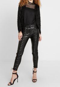 Svart fluffig jacka lager på en genomskinlig svart topp, kombinerad med högmidjade, kroppsnära svarta läderbyxor med sidoremmar och ankelsnören.