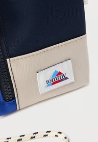 Granatowo-beżowa materiałowa saszetka z widocznym zamkiem błyskawicznym, ozdobiona wielokolorową naszywką z logo "tommy" i teksturowanym plecionym uchwytem.