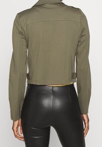 Person wearing a cropped olive green jacket and black high-waisted faux leather pants, shown from the back against a plain background.