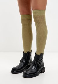 Calcetines de canalé verde olivo hasta la rodilla combinados con botas de cuero negro a la altura del tobillo, que cuentan con un zipper lateral y un detalle decorativo de hebilla.