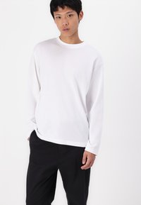 Λευκό βαμβακερό μπλουζάκι με μακριά μανίκια και στρογγυλή λαιμόκοψη, χαλαρή εφαρμογή και κάθοδο στους ώμους, συνδυασμένο με μαύρο παντελόνι. Απαλή υφή.