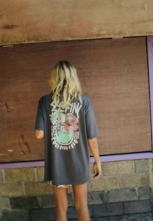 Osoba o blond włosach stoi zwrócona do drewnianej ściany, ubrana w ciemny, oversize'owy T-shirt z kolorową grafiką kwiatową i napisem na plecach.