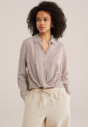 Femme aux cheveux bouclés portant une chemise boutonnée à manches longues rayée marron et blanc, nouée à la taille, et un pantalon beige à cordon de serrage.