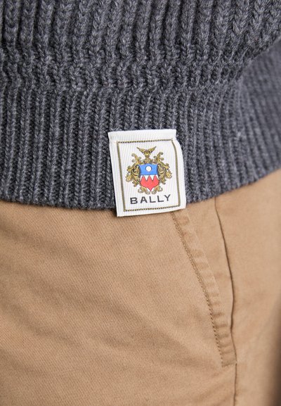 Sivi rebrasti pulover s bijelom etiketom koja prikazuje grb i tekst "BALLY", uparen s kaaki hlačama koje imaju glatku teksturu.
