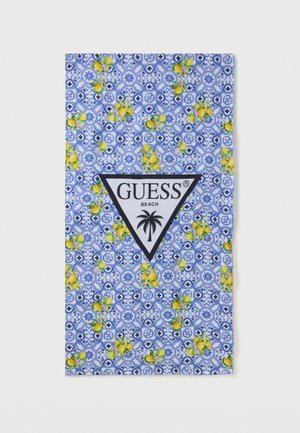 Serviette de plage bleue à motifs, avec des grappes de citrons jaunes et un logo triangulaire blanc central indiquant "GUESS BEACH" avec un palmier noir.