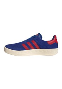 Blue suede sneaker with red stripes, a white rubber sole, and textured accents on the heel. Lace-up design with padded collar.